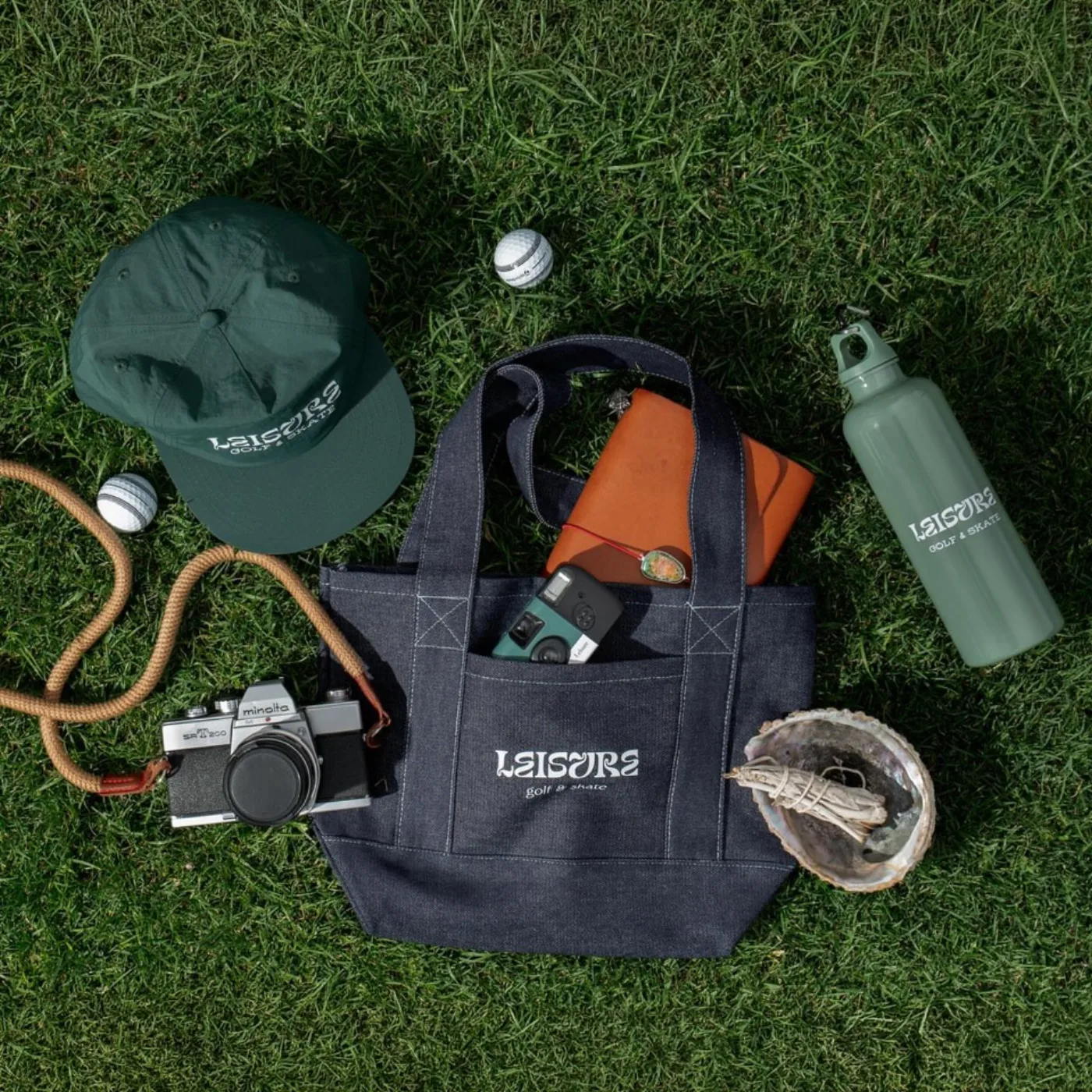 Leisure cap, denim tote, sage bottle and golf balls laid out on grass